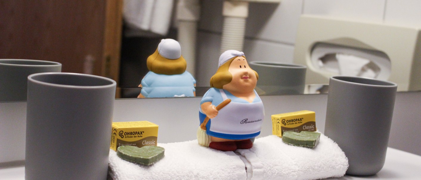 Badezimmer mit zwei grauen Bechern, einer kleinen Figur, Ohrstöpseln und Seife auf einem Handtuch vor einem Spiegel., © TOMAS