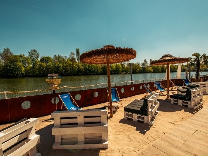 Ein sandiger Strandbereich mit Liegestühlen und Strohdach-Sonnenschirmen am Ufer eines Flusses, umgeben von Bäumen., © Cool-Tours StattReisen Ein sandiger Strandbereich mit Liegestühlen und Strohdach-Sonnenschirmen am Ufer eines Flusses, umgeben von Bäumen., © Cool-Tours StattReisen