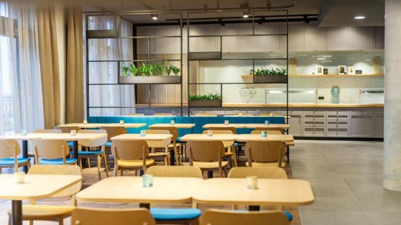 Moderner Speisesaal mit Holztischen und blauen St&uuml;hlen. Im Hintergrund sind Pflanzen und Regale zu sehen. Helle, einladende Atmosph&auml;re., &copy; Arthotel ANA Momentum
