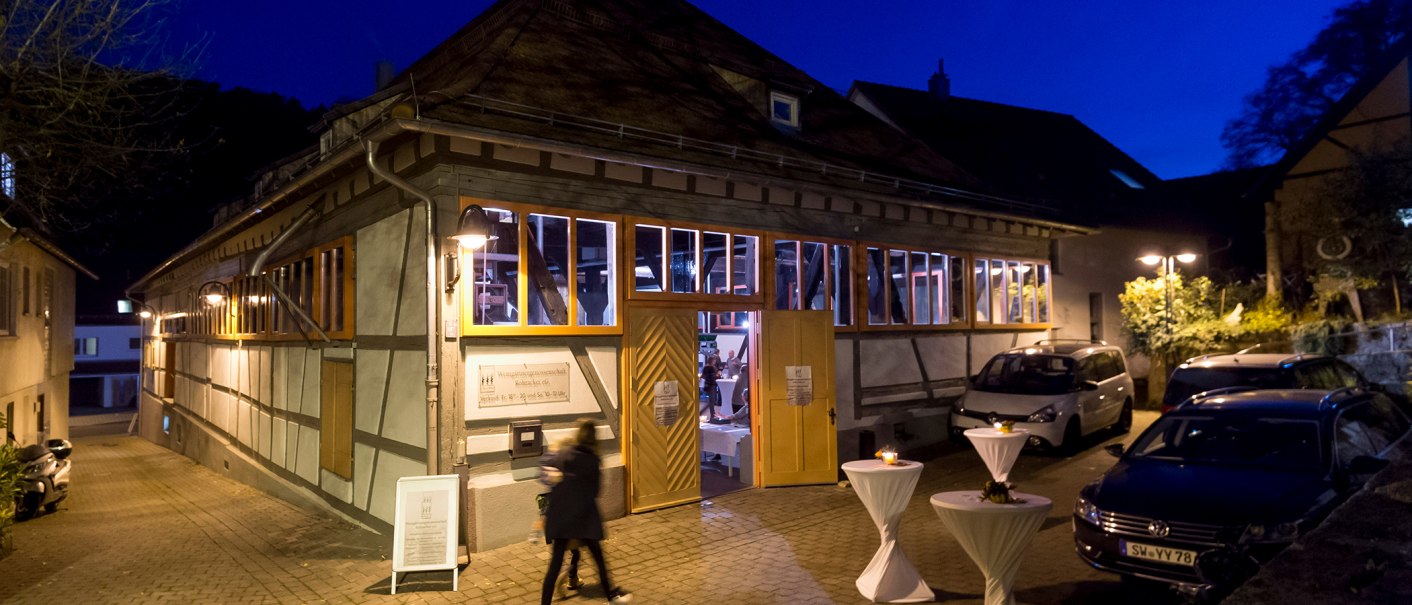 Fachwerkgebäude bei Nacht, beleuchtete Fenster, geöffnete Türen. Davor stehen Stehtische mit weißen Tischdecken. Personen und Autos sind zu sehen., © Steilwerk Rohracker