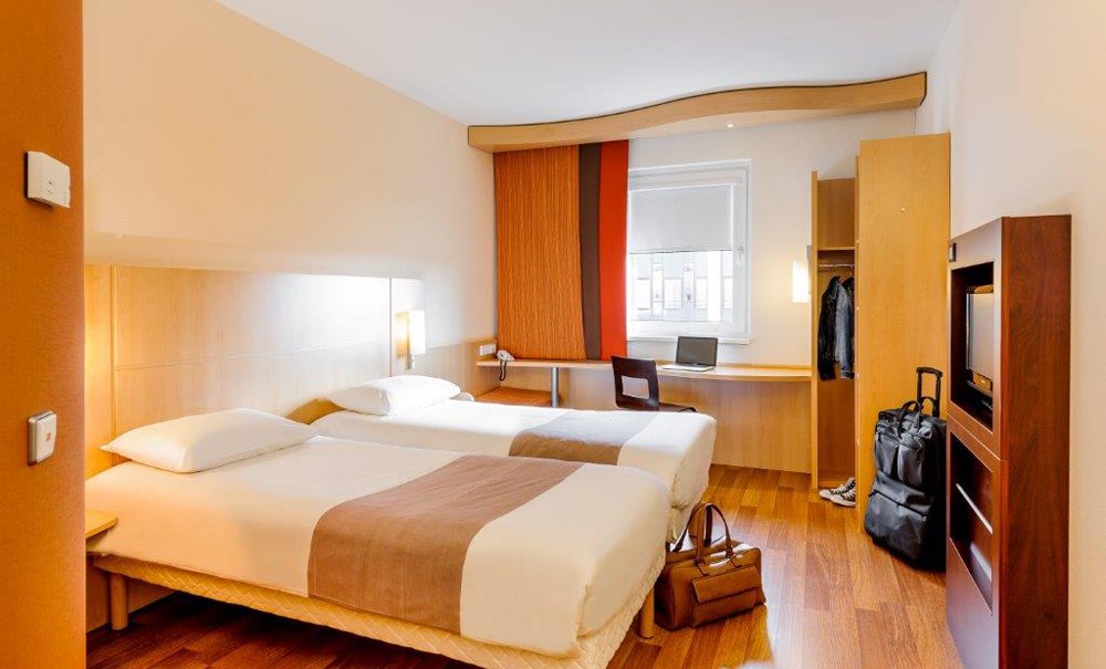 Modernes Doppelzimmer mit zwei Einzelbetten, Schreibtisch, Gepäck und Kleiderschrank. Helle Holzmöbel und warme Farben dominieren den Raum., © Ibis Stuttgart Airport Messe Modernes Doppelzimmer mit zwei Einzelbetten, Schreibtisch, Gepäck und Kleiderschrank. Helle Holzmöbel und warme Farben dominieren den Raum., © Ibis Stuttgart Airport Messe