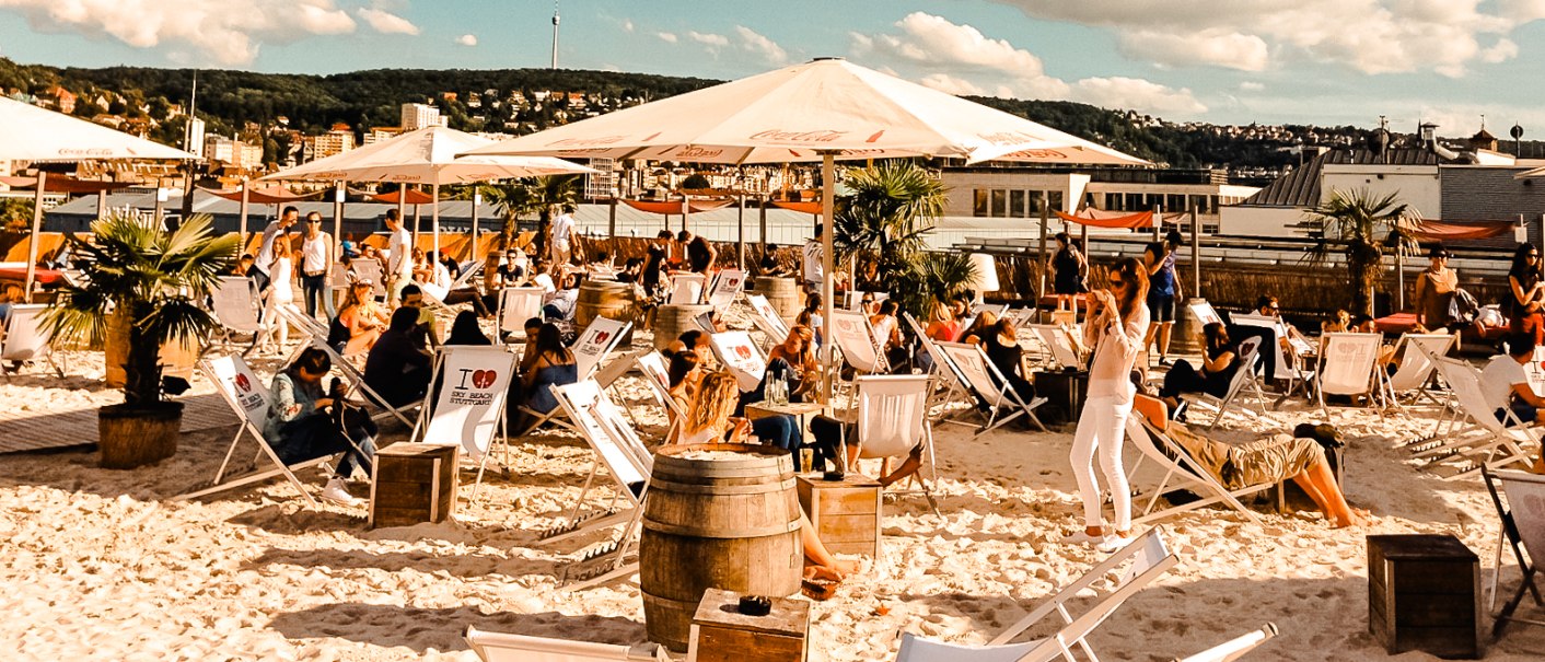 Strandbar mit Liegestühlen und Sonnenschirmen, umgeben von Palmen. Menschen entspannen sich unter blauem Himmel mit Wolken., © Sky Beach