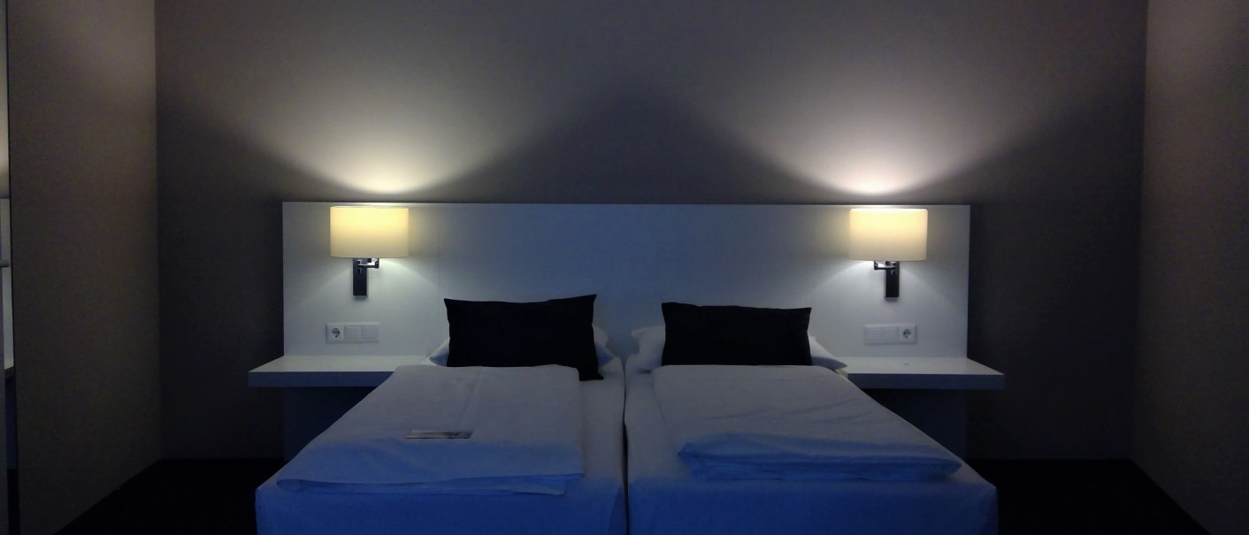 Two single beds with white sheets and black pillows in a hotel room. There are two lamps on the wall that provide warm light., &copy; relexa Waldhotel Schatten