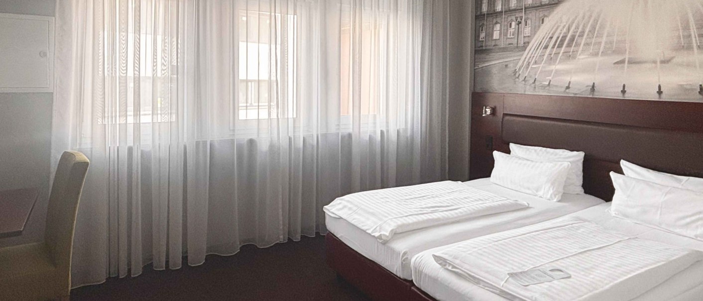 Ein modernes Hotelzimmer mit einem Doppelbett, wei&szlig;en Bettlaken und einem gro&szlig;en Fenster mit transparenten Vorh&auml;ngen. An der Wand ist ein Bild eines Geb&auml;udes mit Brunnen., &copy; Novum Hospitality