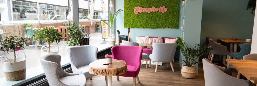 Helles Café mit modernen grauen und pinken Stühlen, Holztischen und grünen Pflanzen. Große Fensterfront und grüne Mooswand mit "Energetic life"-Schriftzug., © SMG