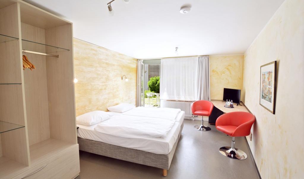 Helles Zimmer mit Doppelbett, zwei roten Stühlen, Schreibtisch, Fernseher und großem Fenster mit Blick ins Grüne., © Abalon Hotel Ideal Helles Zimmer mit Doppelbett, zwei roten Stühlen, Schreibtisch, Fernseher und großem Fenster mit Blick ins Grüne., © Abalon Hotel Ideal