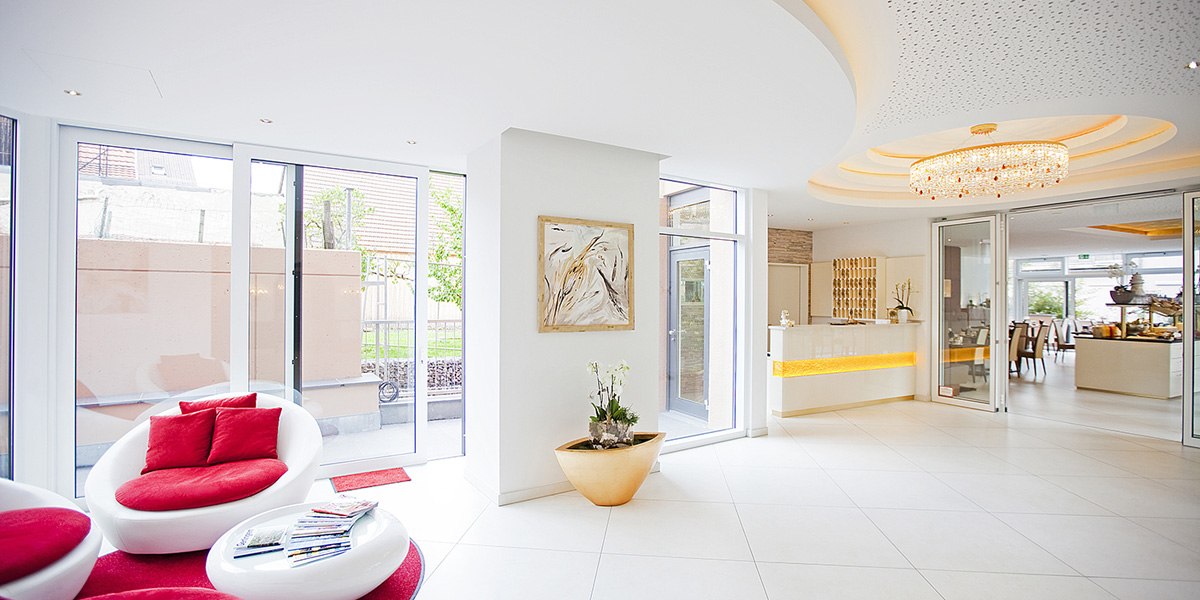 Helle Lobby mit modernen wei&szlig;en M&ouml;beln, roten Kissen, gro&szlig;em Kronleuchter und Glasw&auml;nden. Empfangsbereich mit Pflanzen und Kunstwerk an der Wand., &copy; Hotel Krone