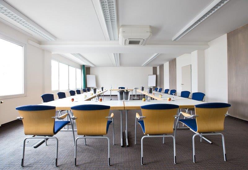 A bright conference room with a U-shaped table arrangement, blue chairs, and flip charts. Large windows let in plenty of light., &copy; bonjour Hotelbetriebs GmbH