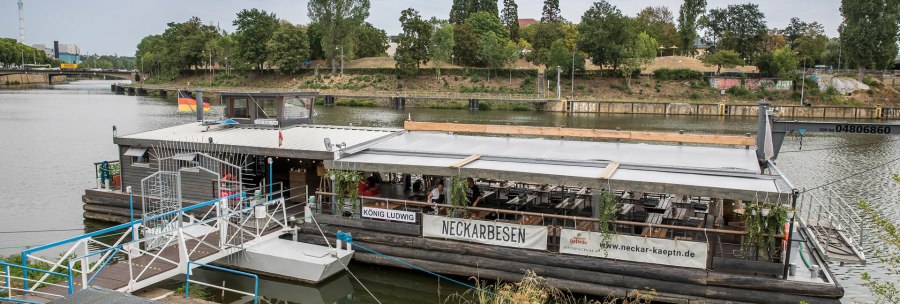 Floating restaurant 'Neckarbesen' on a river, surrounded by trees. A bridge is visible in the background., © all copyrights are reserved by maks richter