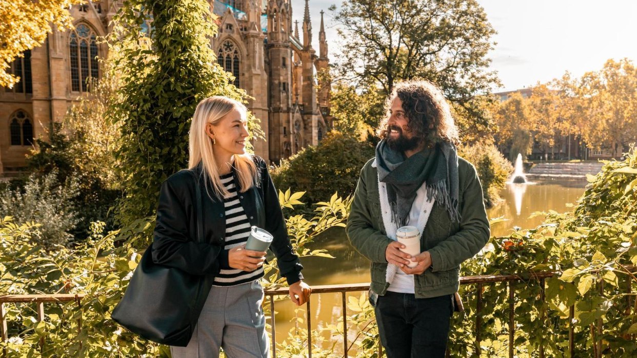 Two people stand smiling in front of a church and a pond, surrounded by autumnal foliage. They are holding coffee cups in their hands., &copy; SMG, Sarah Schmid