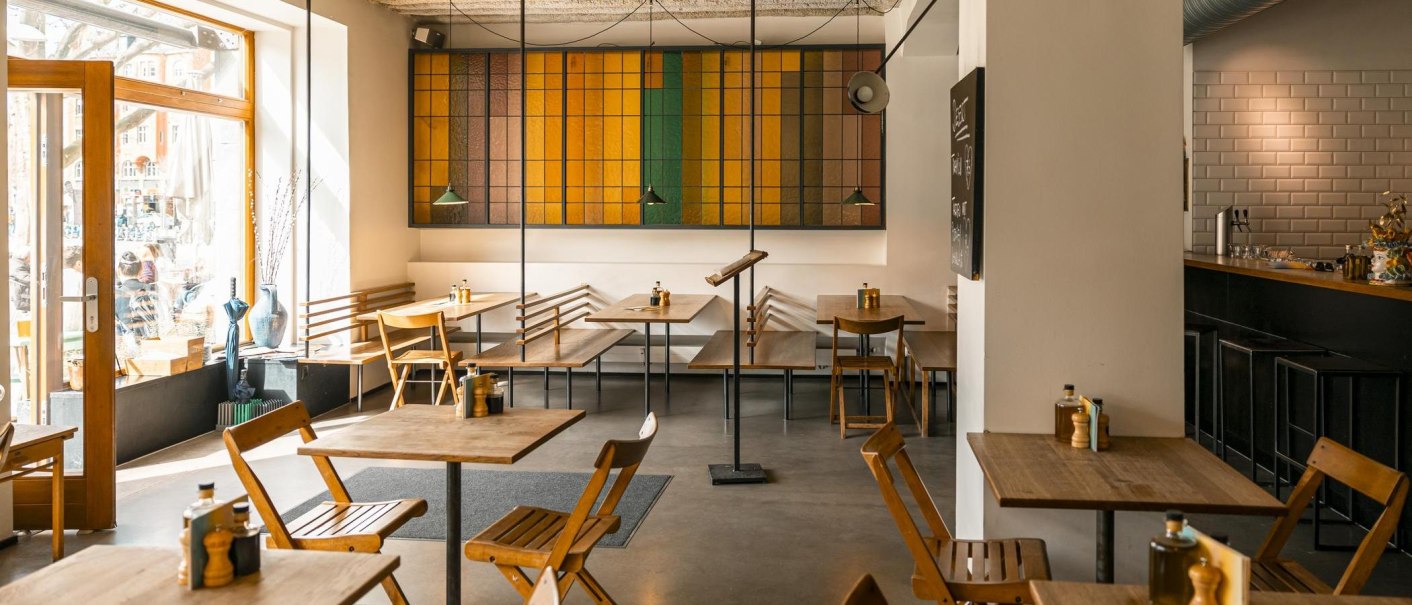 Modernes Café mit Holzmöbeln, buntem Glasfenster und einladender Atmosphäre. Große Fenster lassen viel Licht herein., © SMG, Sarah Schmid