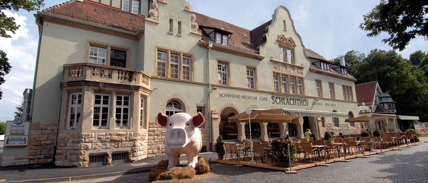 Historic building with pig museum and abattoir café. Large pig figure stands in front of it, surrounded by tables and chairs in the outdoor area., © Buschwerk Mediendesign-Wilhelmer Gastronomie Historic building with pig museum and abattoir café. Large pig figure stands in front of it, surrounded by tables and chairs in the outdoor area., © Buschwerk Mediendesign-Wilhelmer Gastronomie
