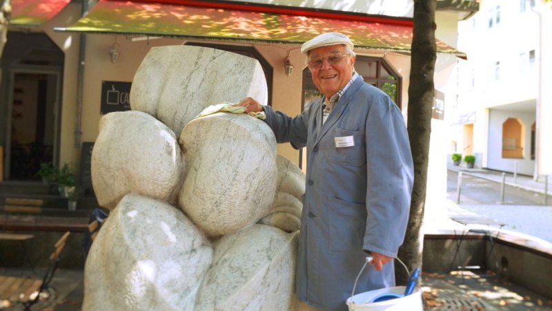 Ein älterer Mann in blauer Arbeitskleidung reinigt eine Steinskulptur. Er trägt eine Mütze und hält einen Eimer in der Hand. Im Hintergrund ist ein Gebäude., © Stuttgart-Marketing GmbH Ein älterer Mann in blauer Arbeitskleidung reinigt eine Steinskulptur. Er trägt eine Mütze und hält einen Eimer in der Hand. Im Hintergrund ist ein Gebäude., © Stuttgart-Marketing GmbH