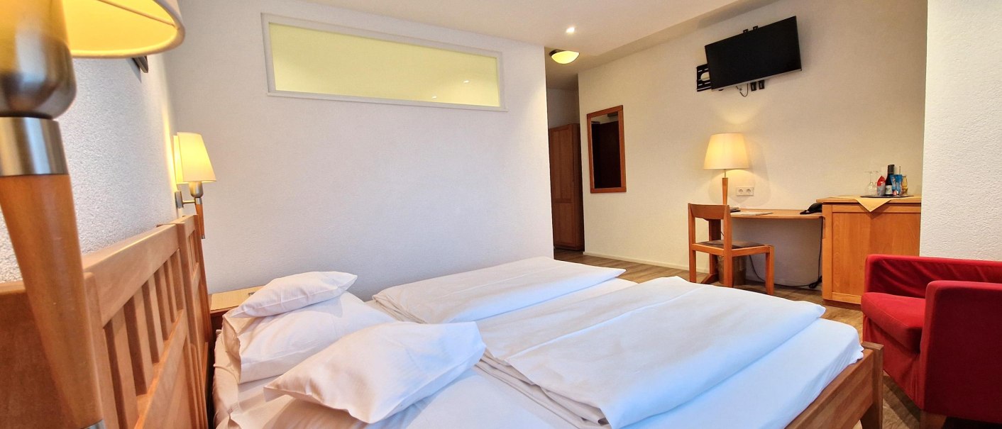 A cozy hotel room with a double bed, a desk, a TV, and a red armchair. Warm lighting and wooden furniture create a welcoming atmosphere., © TOMAS A cozy hotel room with a double bed, a desk, a TV, and a red armchair. Warm lighting and wooden furniture create a welcoming atmosphere., © TOMAS