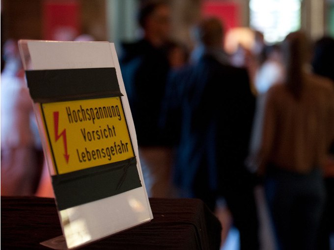 Schild mit Warnung vor Hochspannung, im Vordergrund. Unscharfe Menschenmenge im Hintergrund in einem Innenraum., © Landesmuseum Württemberg Schild mit Warnung vor Hochspannung, im Vordergrund. Unscharfe Menschenmenge im Hintergrund in einem Innenraum., © Landesmuseum Württemberg