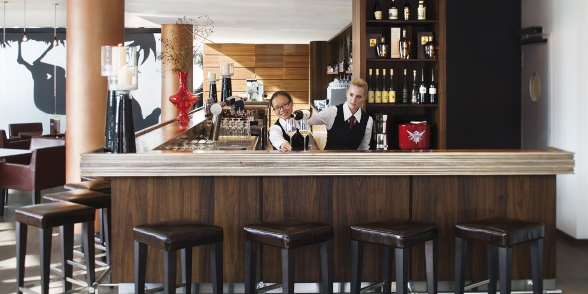 Zwei Barkeeper stehen hinter einer modernen Bar mit Holztheke. Im Hintergrund sind Flaschen und Gläser zu sehen., © Mövenpick Hotel Stuttgart Airport Zwei Barkeeper stehen hinter einer modernen Bar mit Holztheke. Im Hintergrund sind Flaschen und Gläser zu sehen., © Mövenpick Hotel Stuttgart Airport