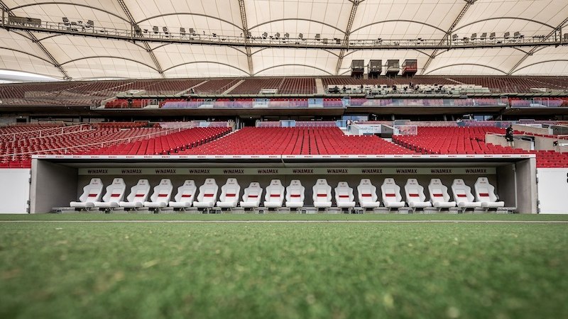 Leere Spielerbank im Stadion mit roten Sitzen und überdachtem Bereich. Der Rasen im Vordergrund und die Tribünen im Hintergrund sind sichtbar., © Mehrwert - BW-Bank Leere Spielerbank im Stadion mit roten Sitzen und überdachtem Bereich. Der Rasen im Vordergrund und die Tribünen im Hintergrund sind sichtbar., © Mehrwert - BW-Bank