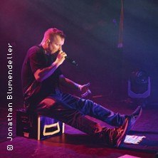 Ein Musiker sitzt auf der B&uuml;hne und singt in ein Mikrofon, umgeben von buntem B&uuml;hnenlicht., &copy; links im Bild