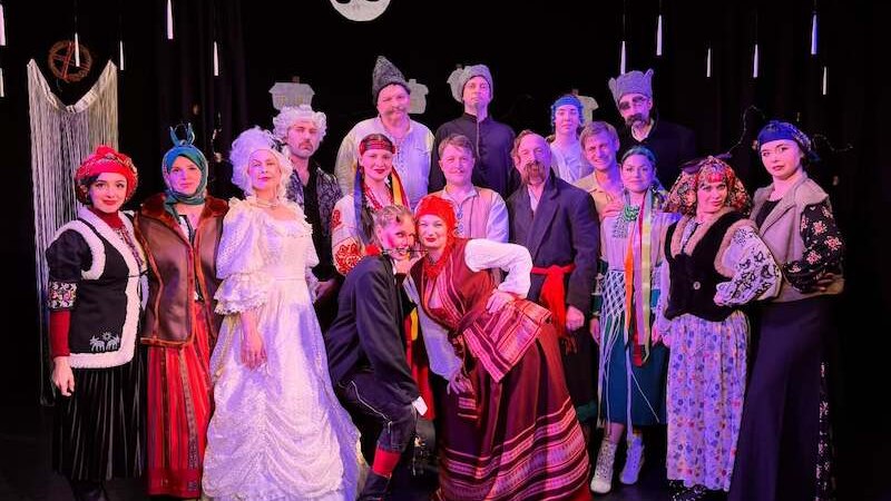 Two people in traditional Ukrainian clothing are standing on a stage, smiling. The man is wearing a cap, and the woman is wearing a red headscarf., © Theater Atelier Two people in traditional Ukrainian clothing are standing on a stage, smiling. The man is wearing a cap, and the woman is wearing a red headscarf., © Theater Atelier