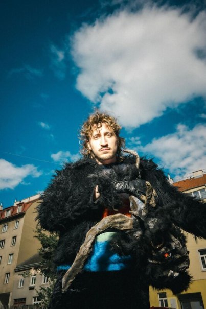A person in a black, fur-like costume with horns stands in front of a blue sky with clouds. Buildings can be seen in the background., &copy; Rosenau Kultur e.V.