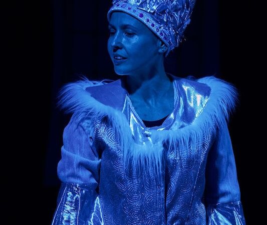 Eine Frau in einem glitzernden Kostüm mit Fellbesatz und einer Krone, beleuchtet von blauem Licht, stellt die Schneekönigin dar., © Theater Atelier Eine Frau in einem glitzernden Kostüm mit Fellbesatz und einer Krone, beleuchtet von blauem Licht, stellt die Schneekönigin dar., © Theater Atelier