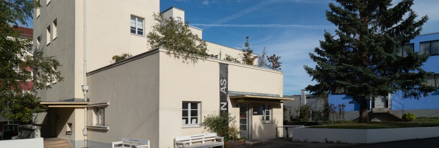 Beige Architekturgalerie mit Flachdach, Bänken und großem Nadelbaum vor blauem Himmel., © Thomas Fütterer Beige Architekturgalerie mit Flachdach, Bänken und großem Nadelbaum vor blauem Himmel., © Thomas Fütterer