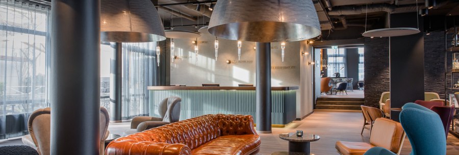 Elegante Hotellobby mit braunem Ledersofa, blauen und grauen Sesseln, großen Hängelampen und einem Empfangstresen im Hintergrund., © TOMAS Elegante Hotellobby mit braunem Ledersofa, blauen und grauen Sesseln, großen Hängelampen und einem Empfangstresen im Hintergrund., © TOMAS