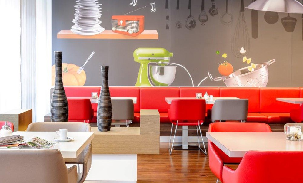 Moderner Frühstücksbereich mit roten und grauen Stühlen, dekorativen Vasen und einer Wand mit Küchenmotiven., © Ibis Stuttgart Airport Messe