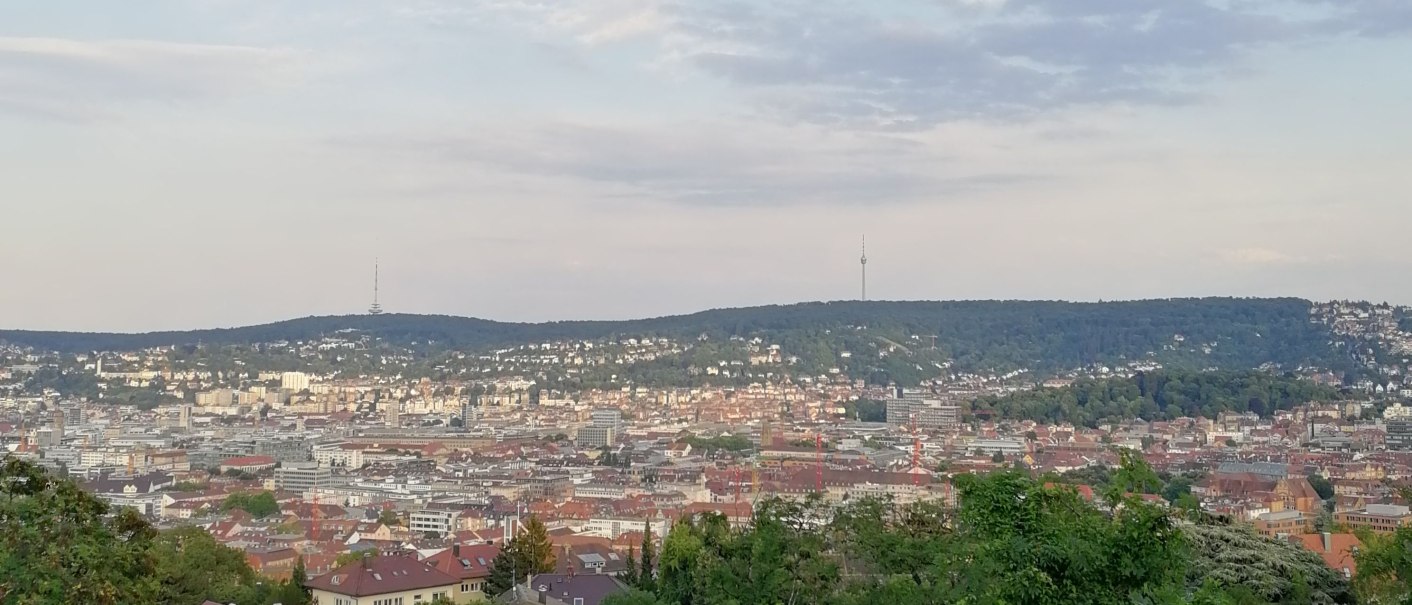 Blick über eine Stadt mit einem Fernsehturm in der Ferne, umgeben von Hügeln und bewaldeten Flächen im Vordergrund., © SMG