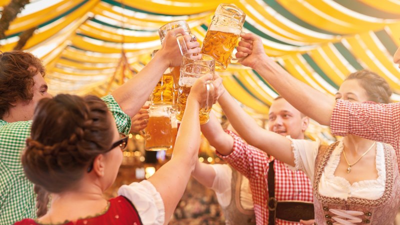 Gruppe in Tracht stößt mit Bierkrügen in einem festlich dekorierten Bierzelt an. Fröhliche Stimmung unter gelb-grünen Stoffbahnen., © SMG, Jean-Claude Winkler