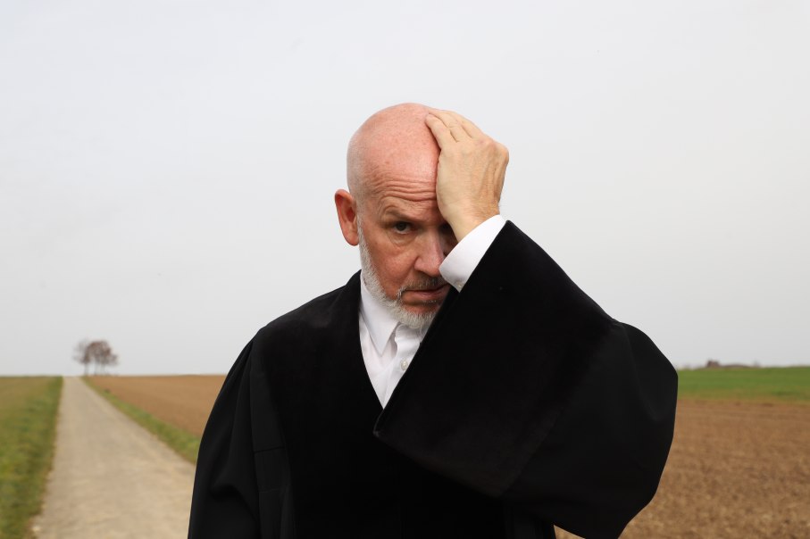 Ein Mann in schwarzer Robe steht auf einem Feldweg und hält sich nachdenklich den Kopf. Der Hintergrund zeigt ein leeres Feld und einen Baum., © Sabine Haymann Ein Mann in schwarzer Robe steht auf einem Feldweg und hält sich nachdenklich den Kopf. Der Hintergrund zeigt ein leeres Feld und einen Baum., © Sabine Haymann