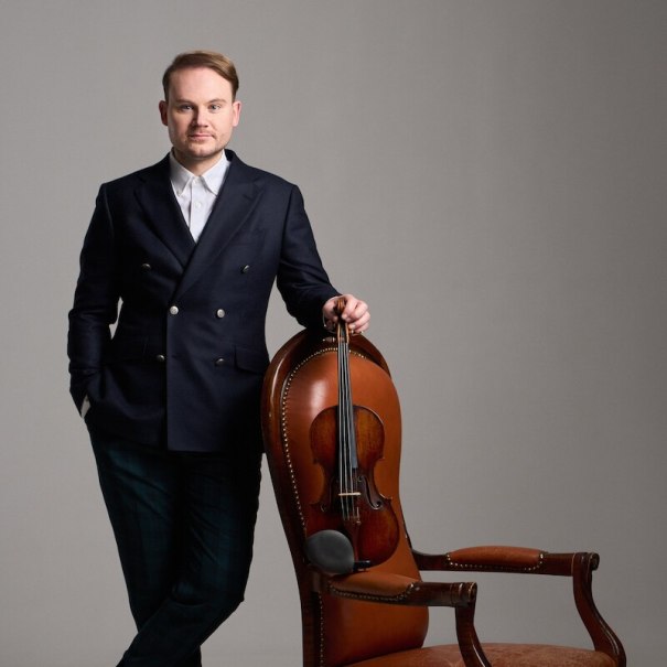 Ein Mann in elegantem Anzug lehnt an einem braunen Lederstuhl, auf dem eine Geige liegt. Der Hintergrund ist neutral grau., © Klassische Philharmonie Bonn e.V. Ein Mann in elegantem Anzug lehnt an einem braunen Lederstuhl, auf dem eine Geige liegt. Der Hintergrund ist neutral grau., © Klassische Philharmonie Bonn e.V.