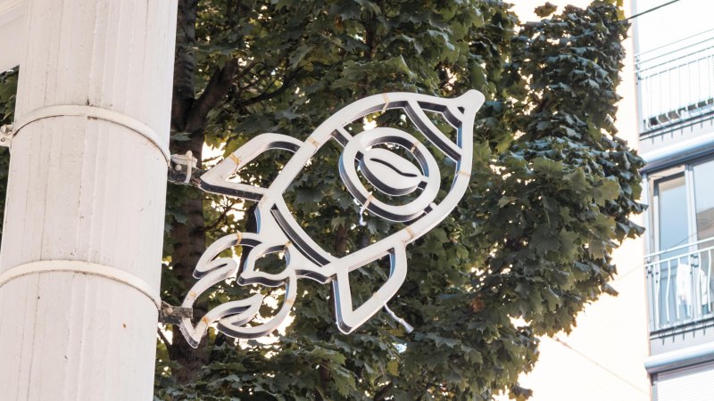 Neonschild in Form einer Rakete mit Kaffeebohne, montiert an einem Baum vor einem Gebäude., © SMG, Sarah Schmid
