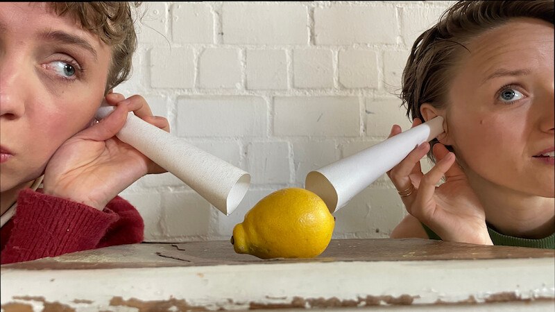 Zwei Personen lauschen mit Papiertrichtern an einer Zitrone auf einem Tisch. Die Szene wirkt humorvoll und kreativ., © FITZ Das Theater animierter Formen Zwei Personen lauschen mit Papiertrichtern an einer Zitrone auf einem Tisch. Die Szene wirkt humorvoll und kreativ., © FITZ Das Theater animierter Formen