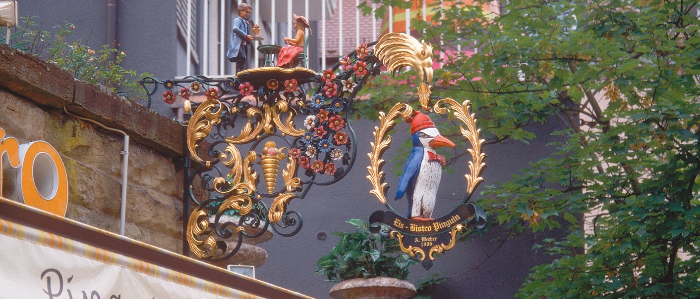 Ein kunstvoll verziertes Schild mit einem Pinguin und Figuren, umgeben von Blumenornamenten, hängt an einer Wand am Eugensplatz., © Stuttgart-Marketing GmbH