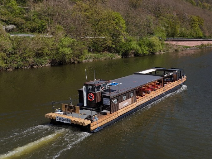 Flussschiff Flunder auf einem Fluss, umgeben von grüner Landschaft. An Bord sind Sitzgelegenheiten für eine BBQ-Tour., © Cool-Tours StattReisen