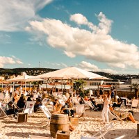 Strandbar mit Liegestühlen und Sonnenschirmen, umgeben von Palmen. Menschen entspannen sich unter blauem Himmel mit Wolken., © Sky Beach
