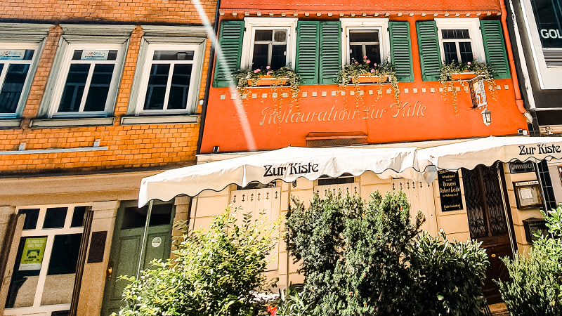 The Zur Kiste wine bar with its red façade, green shutters and awning. Plants adorn the entrance area., © Stuttgart-Marketing GmbH