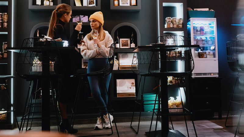 Zwei Frauen in einem stilvollen Café, eine mit gelber Mütze, halten Weingläser. Im Hintergrund sind Regale mit Produkten und ein Kühlschrank zu sehen., © Stuttgart-Marketing GmbH, Alwin Maigler Zwei Frauen in einem stilvollen Café, eine mit gelber Mütze, halten Weingläser. Im Hintergrund sind Regale mit Produkten und ein Kühlschrank zu sehen., © Stuttgart-Marketing GmbH, Alwin Maigler