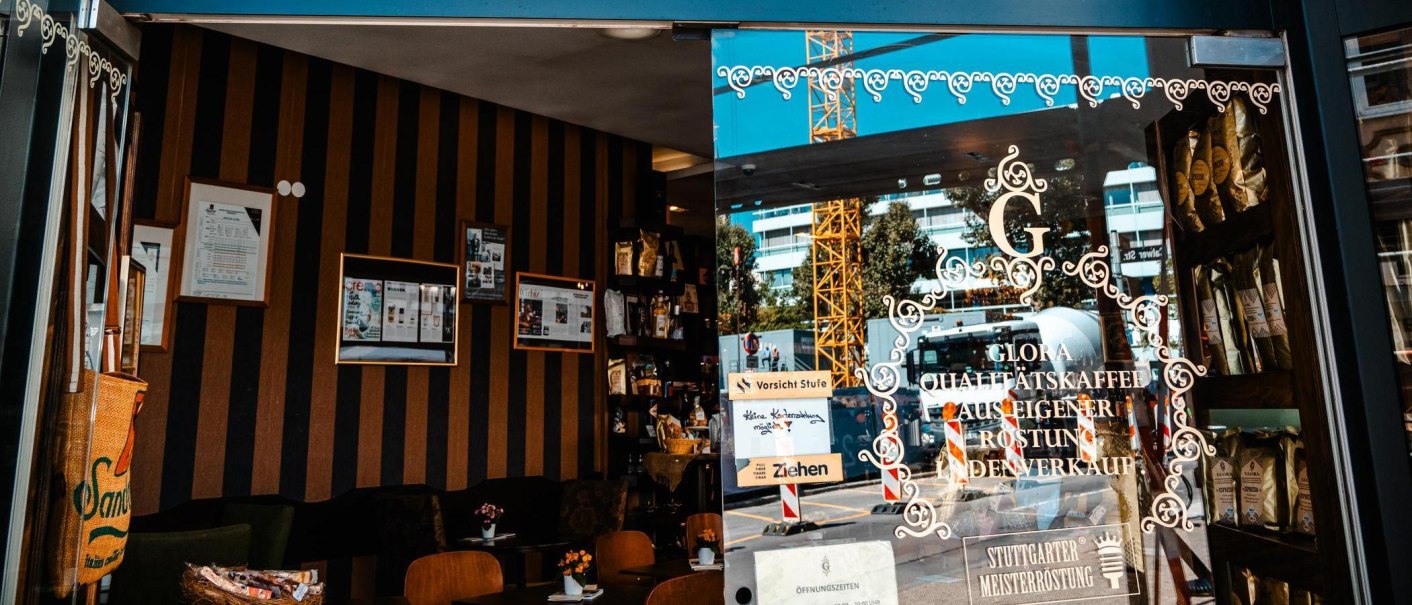 Eingang des Glora Kaffeehauses mit dekorativer Glastür, Innenansicht mit Regalen und Tischen. Draußen ist ein Baukran sichtbar., © Stuttgart-Marketing GmbH, Sarah Schmid