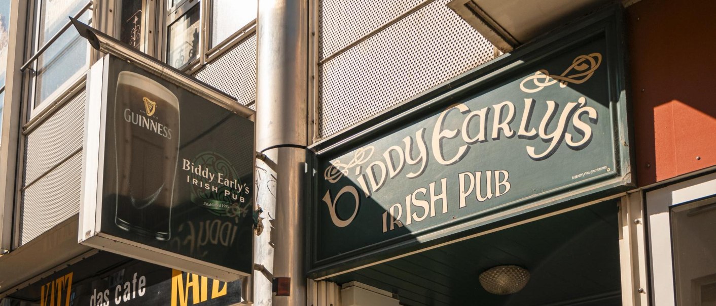 Eingang von Biddy Early's Irish Pub mit grünem Schild und Guinness-Werbung. Sonneneinstrahlung auf der Fassade., © SMG, Sarah Schmid Eingang von Biddy Early's Irish Pub mit grünem Schild und Guinness-Werbung. Sonneneinstrahlung auf der Fassade., © SMG, Sarah Schmid
