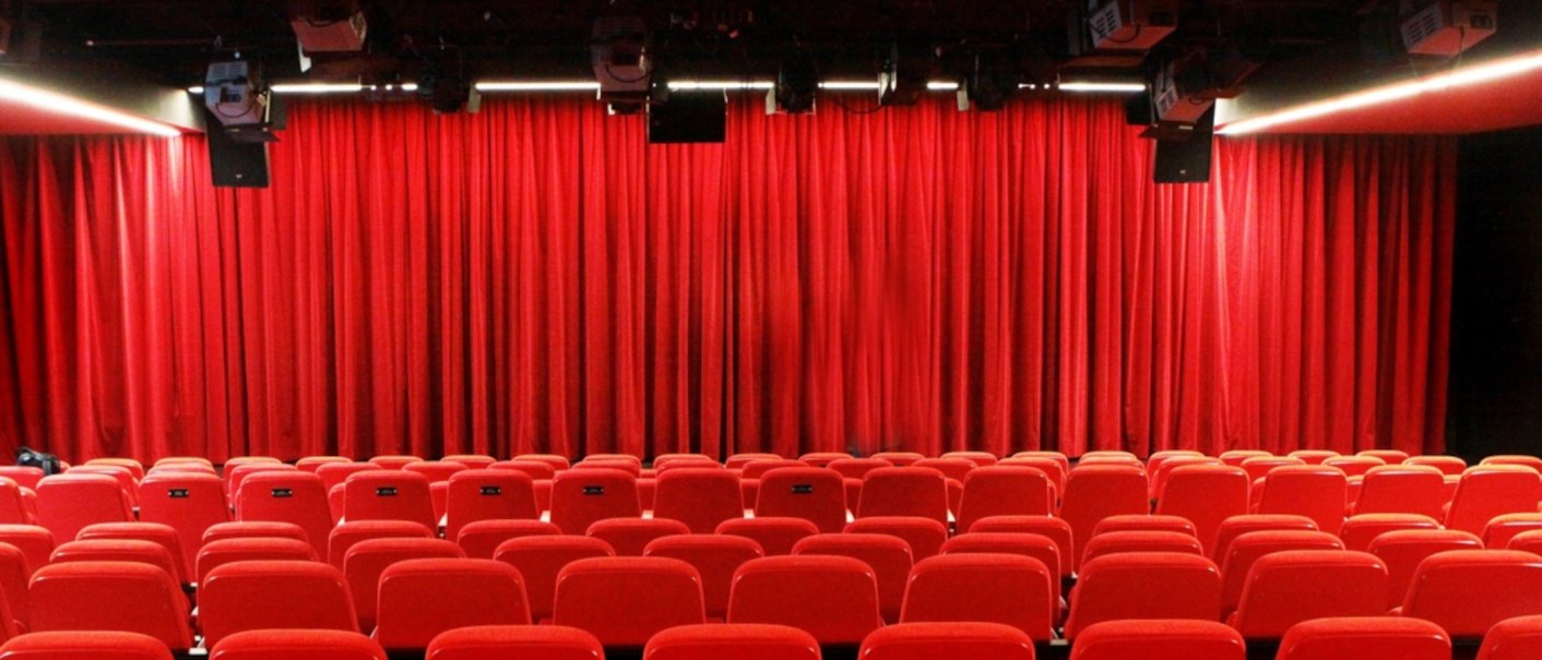 Empty theater hall with red chairs and closed red curtain, lighting and loudspeakers on the ceiling., © Sabine Haymann
