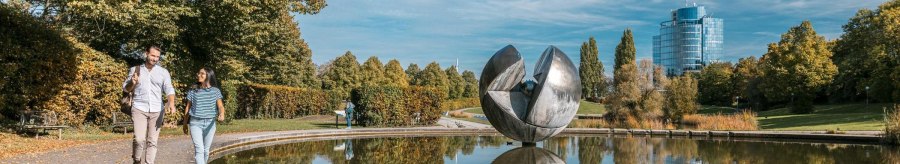 Ein Paar spaziert im Wartbergpark entlang eines Teiches mit einer gro&szlig;en Skulptur und einem modernen Geb&auml;ude im Hintergrund., &copy; Stuttgart-Marketing GmbH, Sarah Schmid