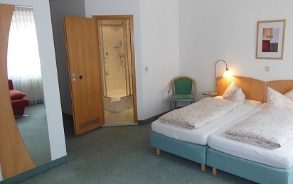 Hotelzimmer mit zwei Einzelbetten, offener Tür zum Bad, Sessel und Wandbild. Helle, schlichte Einrichtung., © TOMAS