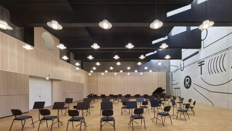 Moderner Saal mit Stühlen und Notenständern, helle Holzverkleidung, geometrische Wandmuster, hängende Lampen., © TOMAS Moderner Saal mit Stühlen und Notenständern, helle Holzverkleidung, geometrische Wandmuster, hängende Lampen., © TOMAS