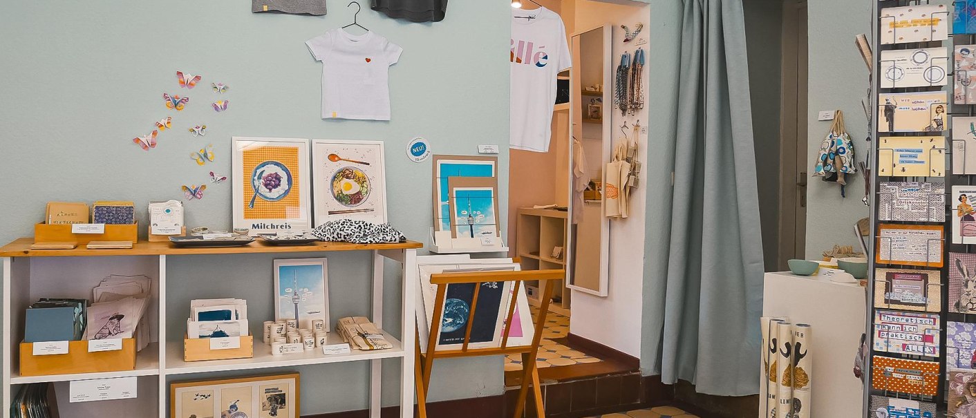 Ein Atelier mit Kunstwerken, T-Shirts an der Wand, bunten Karten und Dekorationen. Regale zeigen verschiedene kreative Produkte., © Vogelsangatelier, Stuttgart