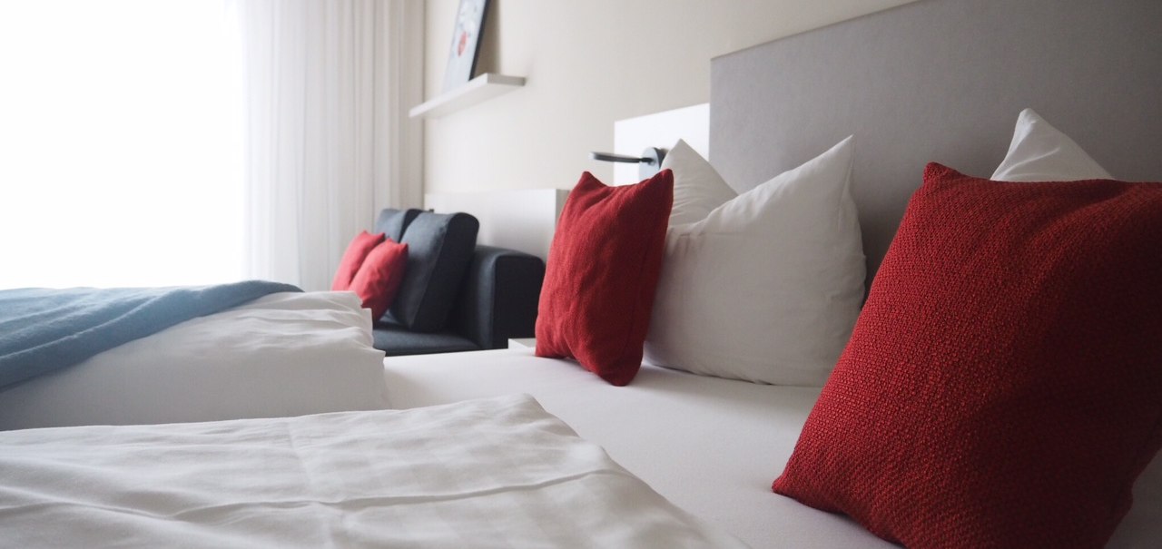 Modern hotel room with white sheets, red cushions and a sofa. A picture hangs on the wall, bright curtains let in light., © TOMAS Modern hotel room with white sheets, red cushions and a sofa. A picture hangs on the wall, bright curtains let in light., © TOMAS