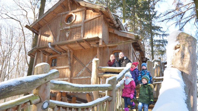 Eine Familie steht auf der Treppe eines verschneiten Baumhauses im Wald. Das Holzhaus ist rustikal und von Bäumen umgeben., © Tripsdrill Natur-Resort GmbH