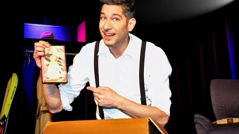 Ein Mann auf einer B&uuml;hne h&auml;lt eine DVD-H&uuml;lle und zeigt darauf. Er tr&auml;gt ein wei&szlig;es Hemd mit Hosentr&auml;gern und l&auml;chelt in die Kamera., &copy; Theaterhaus Stuttgart e.V.