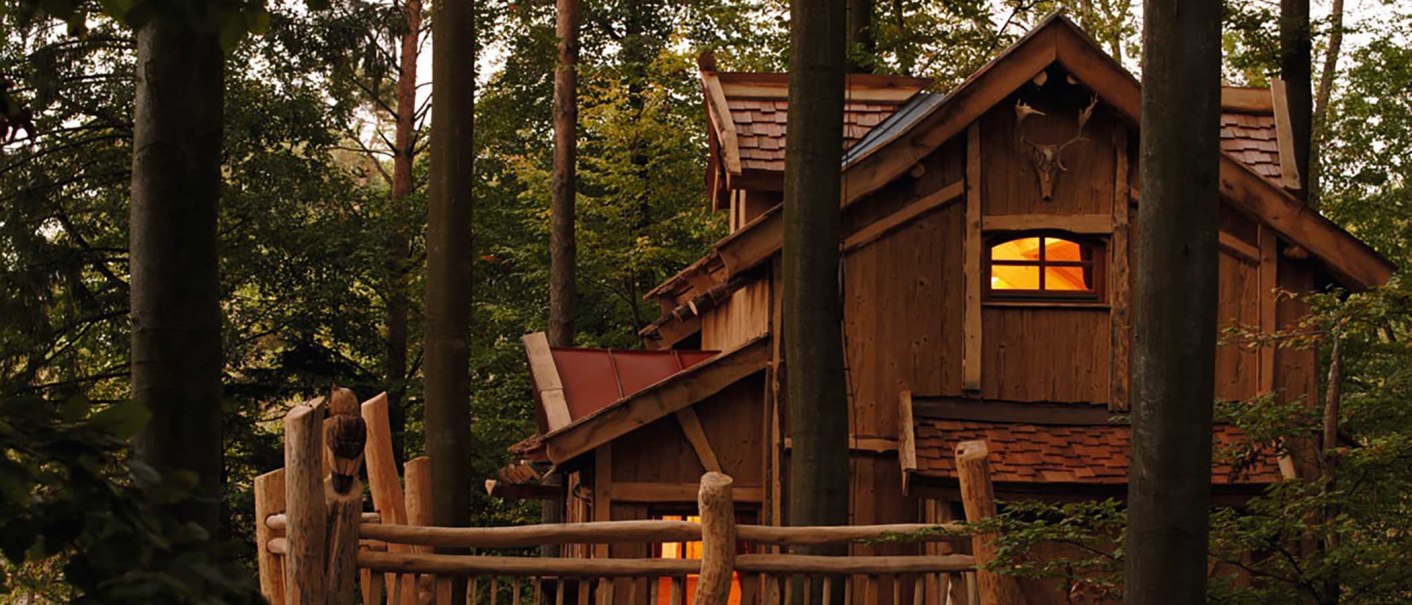 A rustic treehouse in the woods, surrounded by trees. The windows glow warmly, and a wooden fence surrounds the house., &copy; Tripsdrill Natur-Resort GmbH